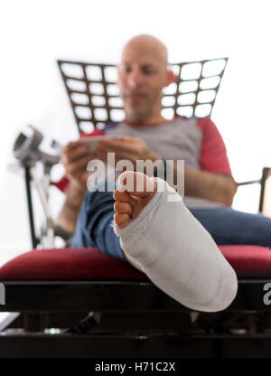 Young man with a broken ankle and a white cast on his leg, sitting on a couch with crutches nearby, surfing the web on his phone Stock Photo