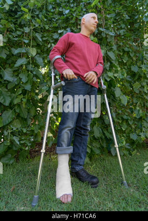 Young man with a broken ankle and a white fiberglass and plaster cast on his leg, getting some fresh air in the garden while wal Stock Photo
