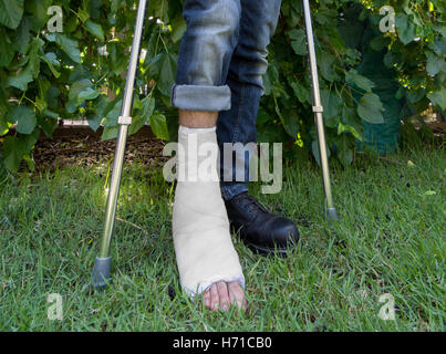 Young man with a broken ankle and a white fiberglass and plaster cast on his leg, getting some fresh air in the garden while wal Stock Photo