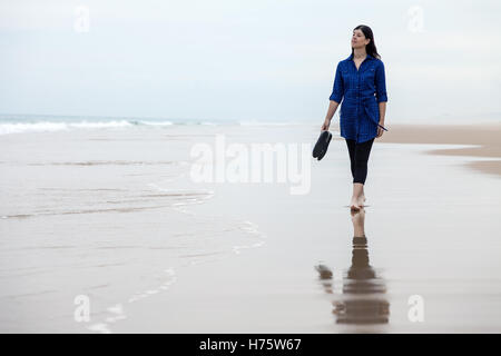 Young woman walking alone in a deserted beach reflected on the wet sand on an Autumn day. Stock Photo