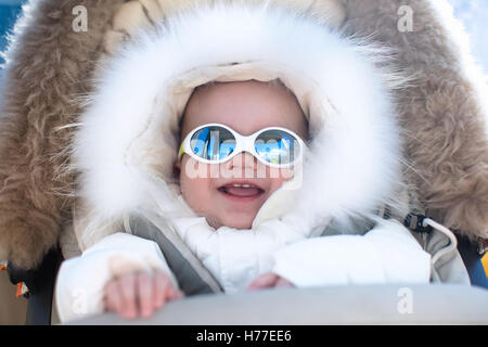Little baby in warm stroller wearing fur hood snow suit enjoying winter ski vacation in alpine resort. Sunglasses for infants Stock Photo