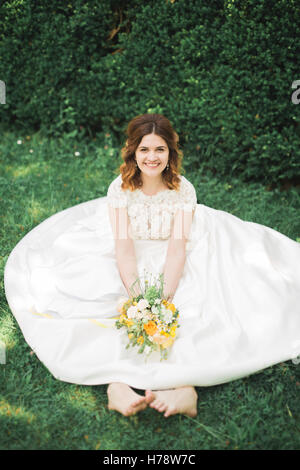 Lovely bride sitting on ground holding a bouquet smiling at camera Stock Photo
