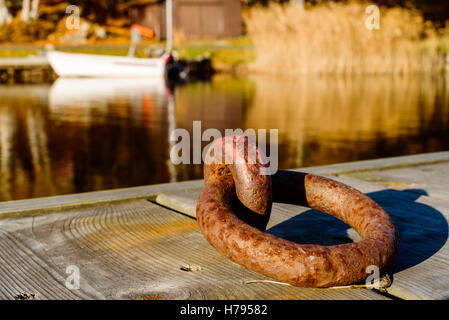 Rusty old mooring ring on wooden pier with boat and coastline in blurred background. Stock Photo