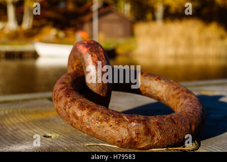 Rusty old mooring ring on wooden pier with boat and coastline in blurred background. Stock Photo