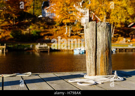 Old gnarled wooden bollard on a pier. Loose white rope lying twirled at the base. Water and opposite coastline in background. Stock Photo