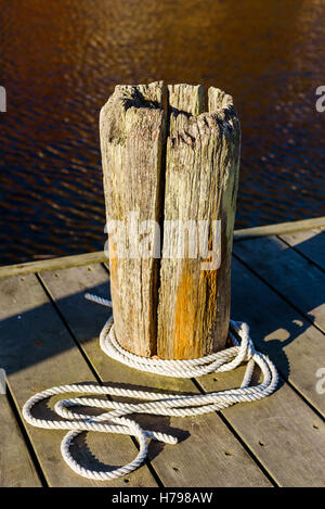 Old gnarled wooden bollard on a pier. Loose white rope lying twirled at the base. Water in background. Stock Photo