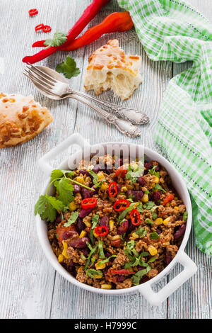 Chili con carne in a white casserole on wooden table with fresh vegetables and bread. Cooked with ground beef, tomatoes, peppers Stock Photo