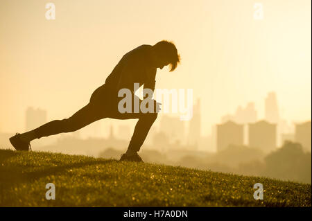 Male jogger stretching on the grassy top of Primrose Hill in front of a misty golden sunrise view of the London city skyline Stock Photo