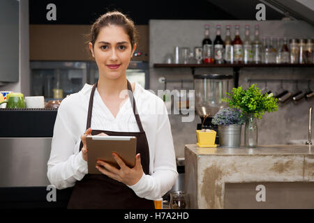 Beautiful Caucasian female barista using tablet and smiling inside coffee shop Stock Photo
