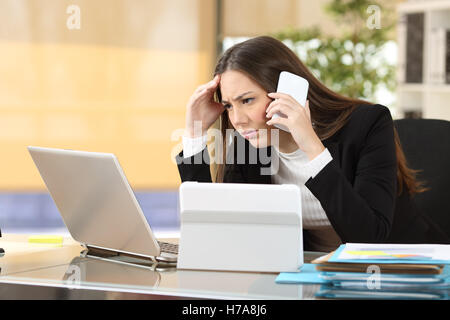 Worried executive talking on the phone trying to solve problems with multiple devices sitting in a desktop at office Stock Photo