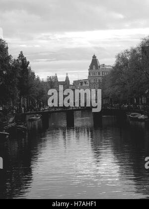Black and white canal in Amsterdam in autumn. Central view of a water canal with a bridge and towers in the distance. Stock Photo