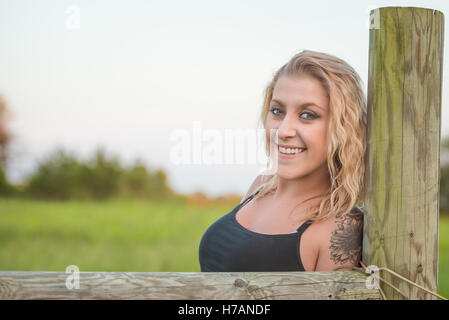 Beautiful blonde woman with tattoo leaning on fence post Stock Photo