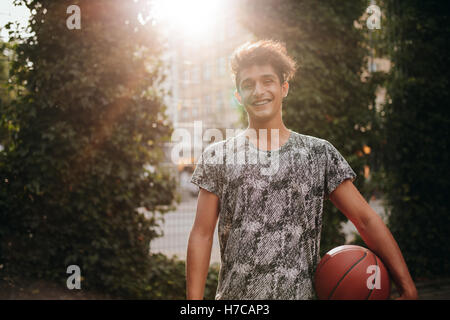 Portrait of handsome young basketball player holding a ball on outdoor court. Smiling teenage streetball player looking at camer Stock Photo