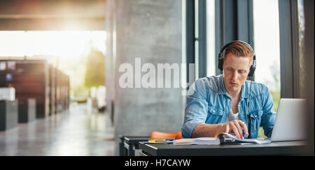 Portrait of young student wearing headphones sitting at the table in library and reading book. University student finding inform Stock Photo
