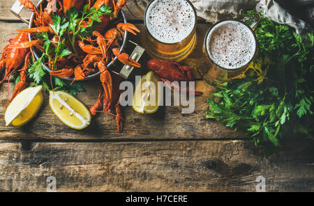 Two mugs of wheat beer and boiled crayfish in pan served with with lemon and parsley over rustic wooden background, top view, co Stock Photo