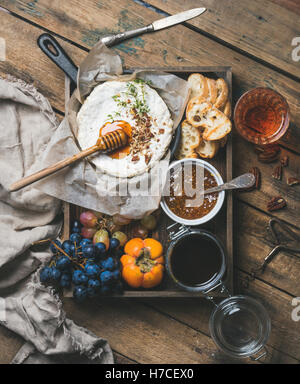 Cheese, fruit and wine set. Camembert in small pan with honey, nuts and herbs, grapes, persimmon, fig jam, baguette slices and g Stock Photo