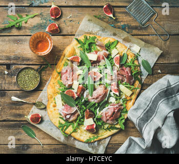 Fig, prosciutto, arugula and sage flatbread pizza with glass of rose wine on wax paper over rustic wooden background, top view Stock Photo