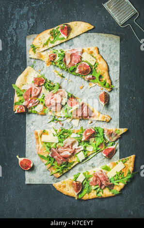 Fig, prosciutto, arugula and sage flatbread pizza cut into pieces on baking paper over black grunge stone background, top view, Stock Photo