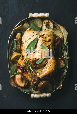 Oven roasted whole chicken with onion, apples and sage in metal serving tray over dark stone background, top view. Stock Photo