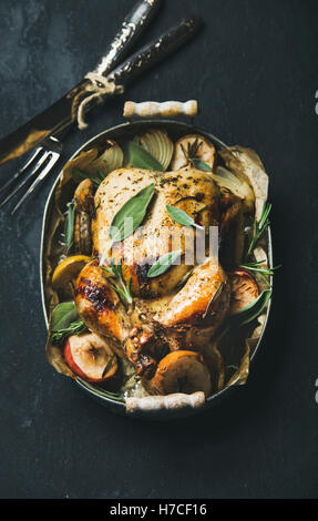 Oven roasted whole chicken with onion, apples and sage in serving tray with cutlery over dark stone background, top view, select
