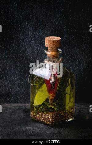 Glass bottle of spicy olive oil with rosemary, red hot chili peppers and bay leaf standing over black textural background. Medit Stock Photo