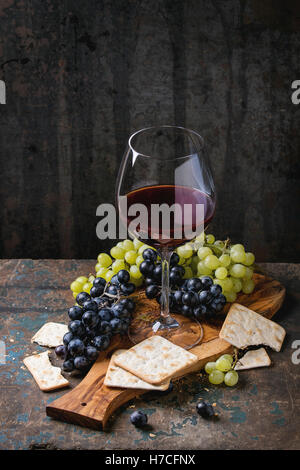 Bunches of ripe wet red and white grapes with crackers snack and glass of red wine on olive wood cutting board over old dark woo Stock Photo