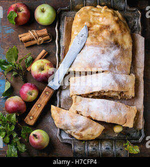 Sliced homemade apple strudel served with fresh apples with leaves, cinnamon sticks and sugar powder on vintage metal tray with Stock Photo
