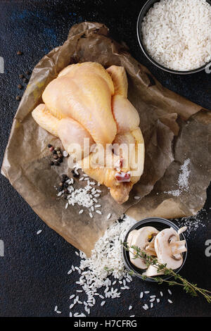Whole raw mini chicken on baking paper with seasoning, thyme, sliced mushrooms and bowl of uncooked white rice over black textur Stock Photo