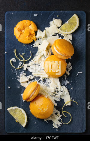 Whole and broken orange lemon homemade macaroons with chopped white chocolate and citrus sugar and zest on dark glass board over Stock Photo