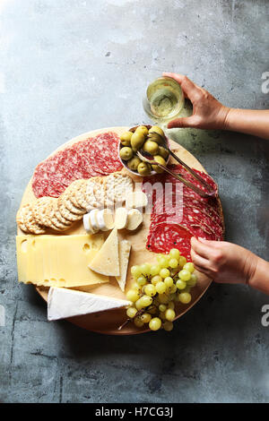 Cheese board with assorted sausages and salami, fresh fruit and various cheeses.Female hand reaching salami from a platter. Stock Photo