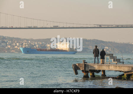 People watch a cargo ship sail under the Bosphorus bridge, from a pier on the Asian Side. Stock Photo