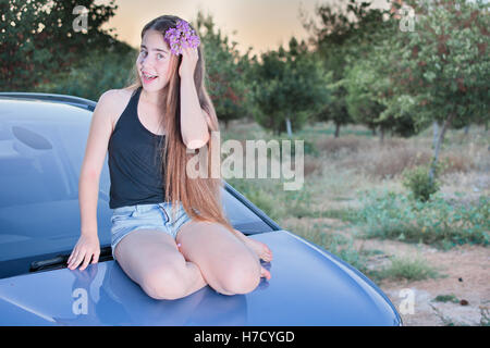 A 13 year old teenage girl with braces on her teeth sitting on a car with flowers in her long hair enjoying the summer sunset Stock Photo