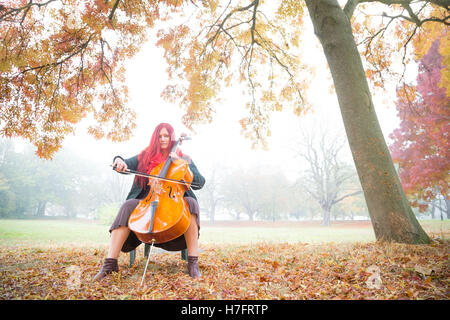 Portrait of a redheaded women posing with a cello under Autumn trees and brown/orange foliage in a west London park, UK. Stock Photo