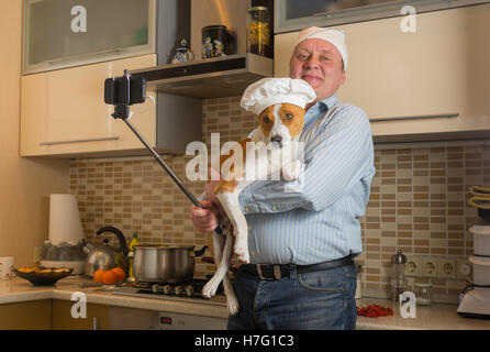 Funny mature man doing selfie in a kitchen with cute basenji dog wearing white cap Stock Photo