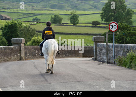 Rear view of young female, riding a grey horse on a quiet, scenic, country road - Burnsall village, Yorkshire Dales England. Stock Photo