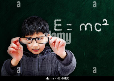 Smart Asian boy holding up eyeglasses on chalkboard background with mathematical equation and copy space. Stock Photo