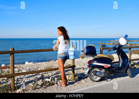 Woman gazing out at the sea next to Vespa Primavera scooter, Marina di Montemarciano, Indian, Alaska Marche, Italy Stock Photo