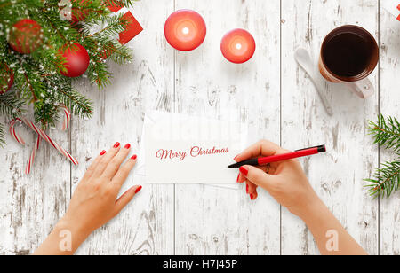 Woman write Christmas greeting message on paper. Top view of white wooden table with christmas tree, decorations, candle, tea Stock Photo