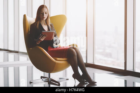 Young serious successful woman entrepreneur in a red dress and jacket sitting on yellow armchair with tablet Stock Photo