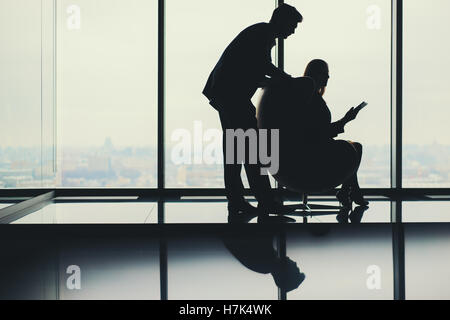 Two silhouettes: businessman standing near his female colleague with tablet sitting on armchair next to huge window in office Stock Photo