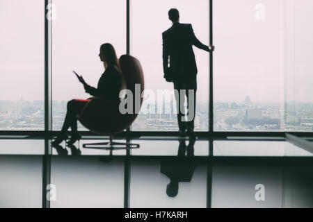 Blurred silhouettes of businessman standing next to big window, looking at cityscape outside, his female colleague with tablet Stock Photo