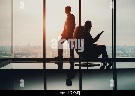 Silhouette of businessman standing next to big window, looking at blurred cityscape outside, his female colleague with tablet Stock Photo