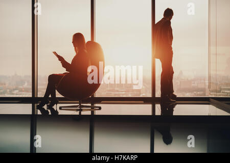 Silhouette of businessman standing next to window, looking at blurred city outside on sunset, his female colleague with tablet Stock Photo