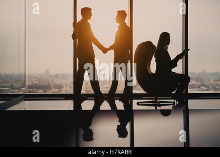 Silhouettes of two young businessmen in luxury office interior completing deal with handshake, female secretary with tablet Stock Photo
