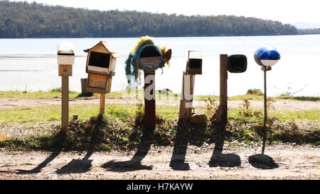 Six letter boxes for different addresses, in a country town. They are casting shadows on the ground, with a rural lake backdrop. Stock Photo
