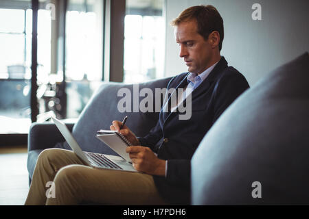 Businessman sitting on the sofa with laptop and writing notes in diary Stock Photo