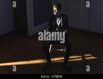 A young man wearing a black leather jacket sits on a chair in an empty room looking off to the side; Tarifa, Cadiz, Andalusia, Spain Stock Photo