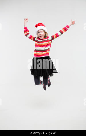 A young girl wearing a red sweater and santa hat, jumping in the air with joy, arms outstretched, against a white background Stock Photo