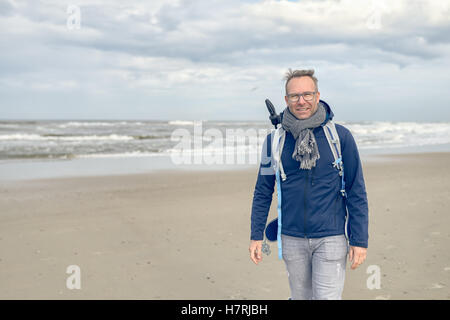 Middle-aged man wearing glasses, a backpack and a scarf walking along a cold blustery beach on a rainy overcast autumn or winter Stock Photo
