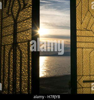 Sunlight glowing at sunset and reflected in the tranquil ocean water as viewed through a window with gold and black shutters Stock Photo